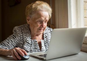 An elderly woman sitting at the table and types on laptop.